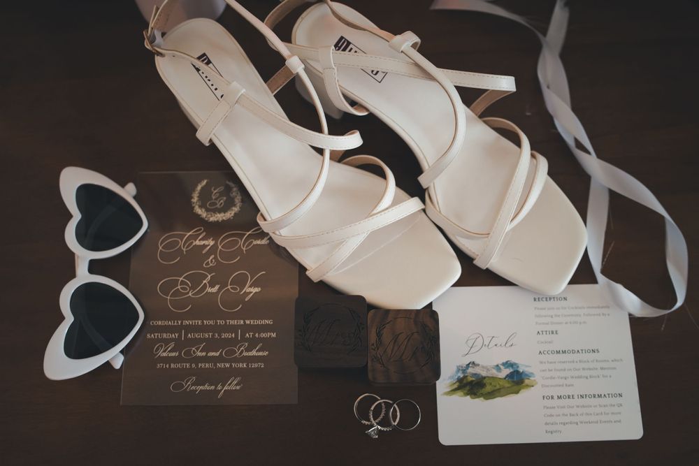 Wedding details including elegant white bridal sandals, heart-shaped sunglasses, and wedding invitation cards on dark surface.