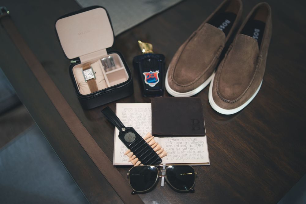 Wedding accessories including brown loafers, sunglasses, and jewelry box arranged on dark wooden surface.