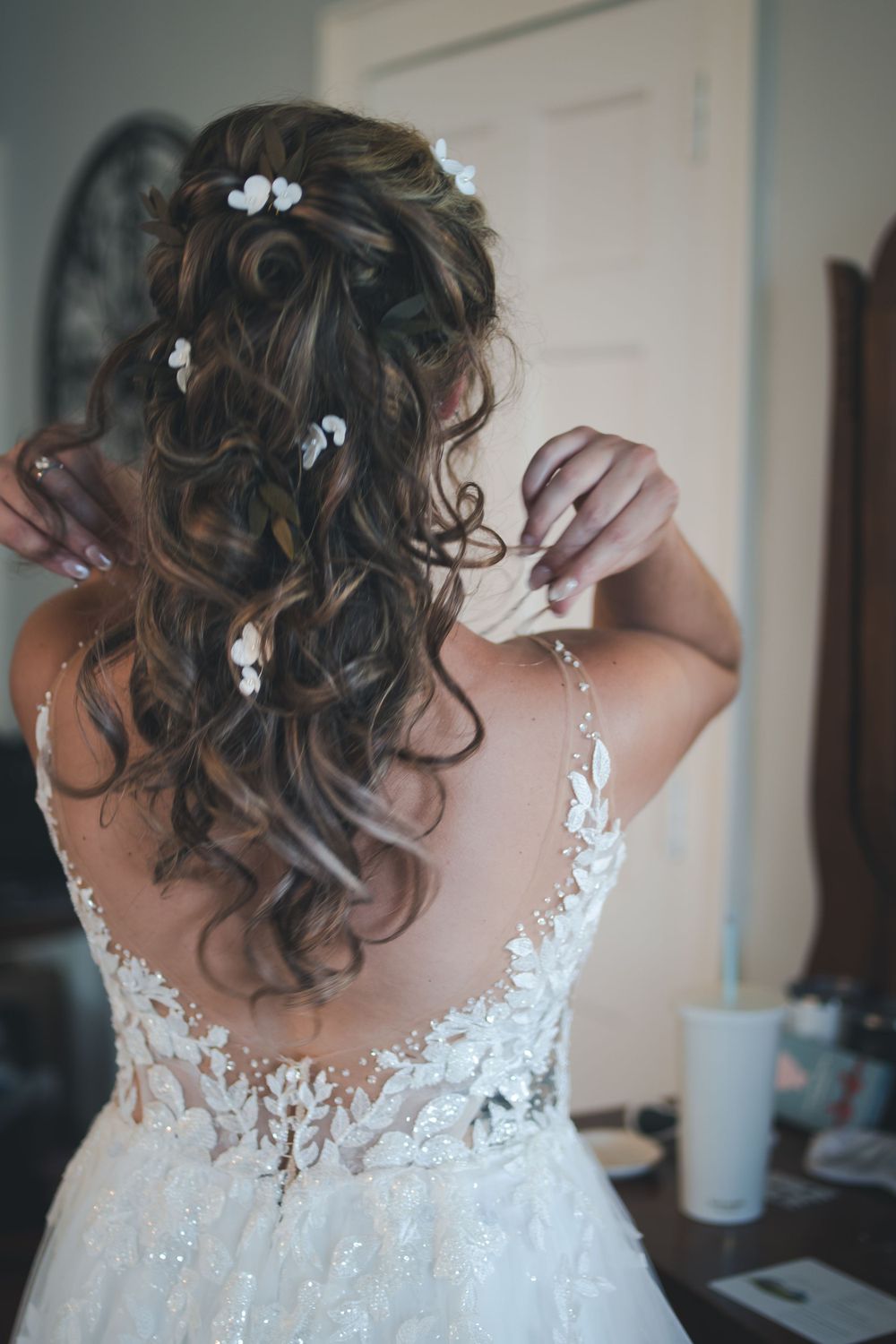Back view of intricate wedding dress with lace detail and curled hairstyle adorned with small white flowers.