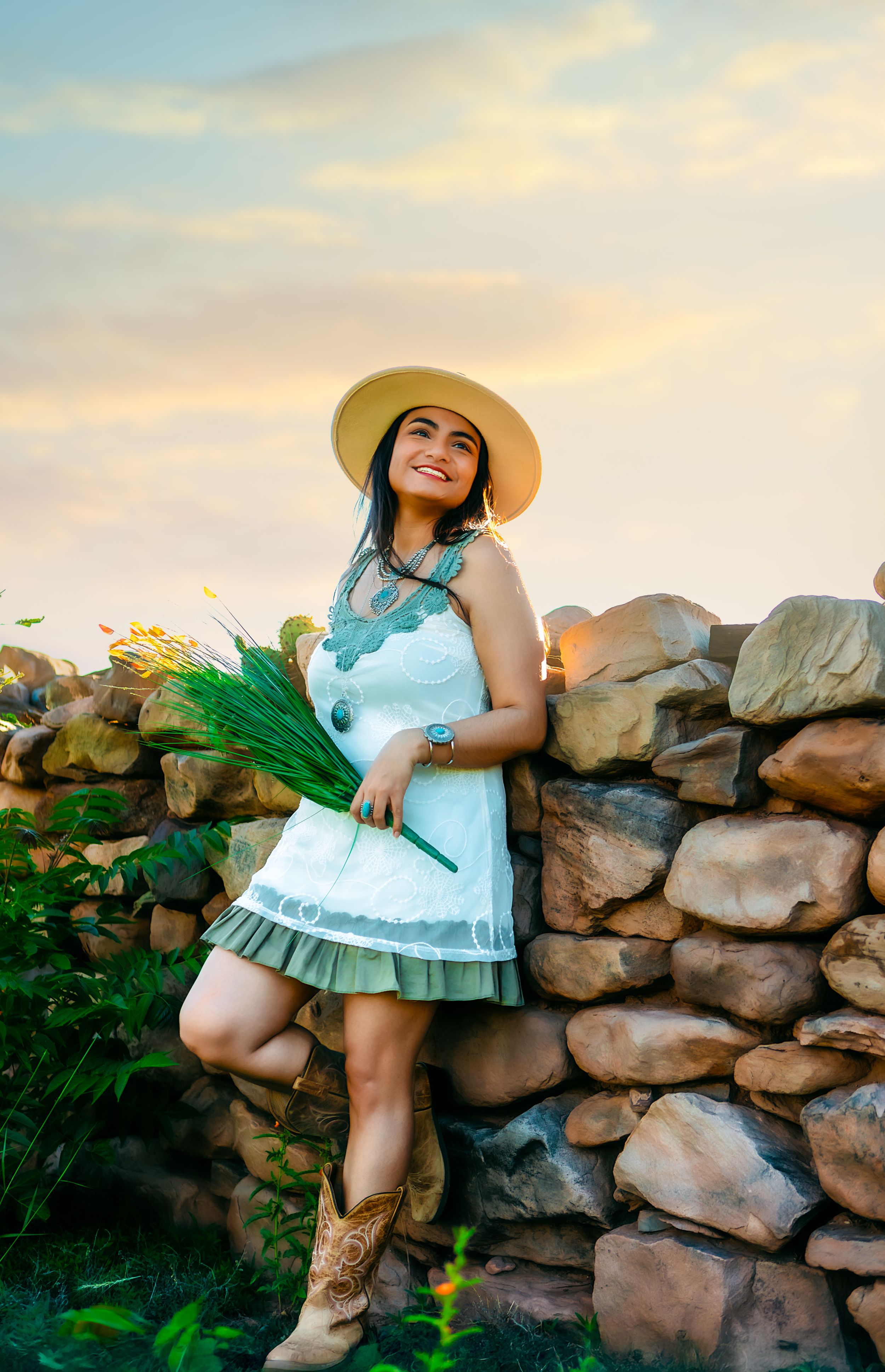 Model in white dress and sunhat perches on rustic stone wall during golden hour photoshoot.