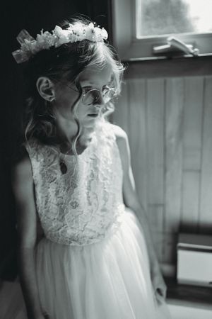 Young girl in white dress and floral crown poses by window in black and white.