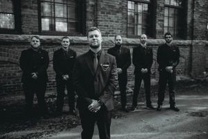 Groomsmen posed in black attire against industrial brick building in monochrome style.