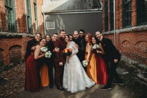 Wedding party in burgundy and yellow dresses gather outdoors against rustic brick walls.