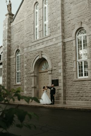 A couple in wedding attire stands at the entrance of a stone church with tall arched windows and doorway.