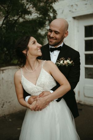A tender moment between newlyweds sharing an embrace against a white exterior wall.
