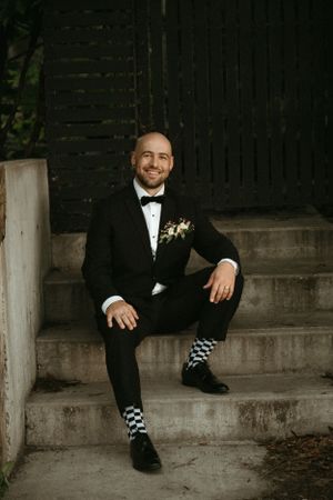 A groom in a tuxedo and checkered socks sits on concrete steps.