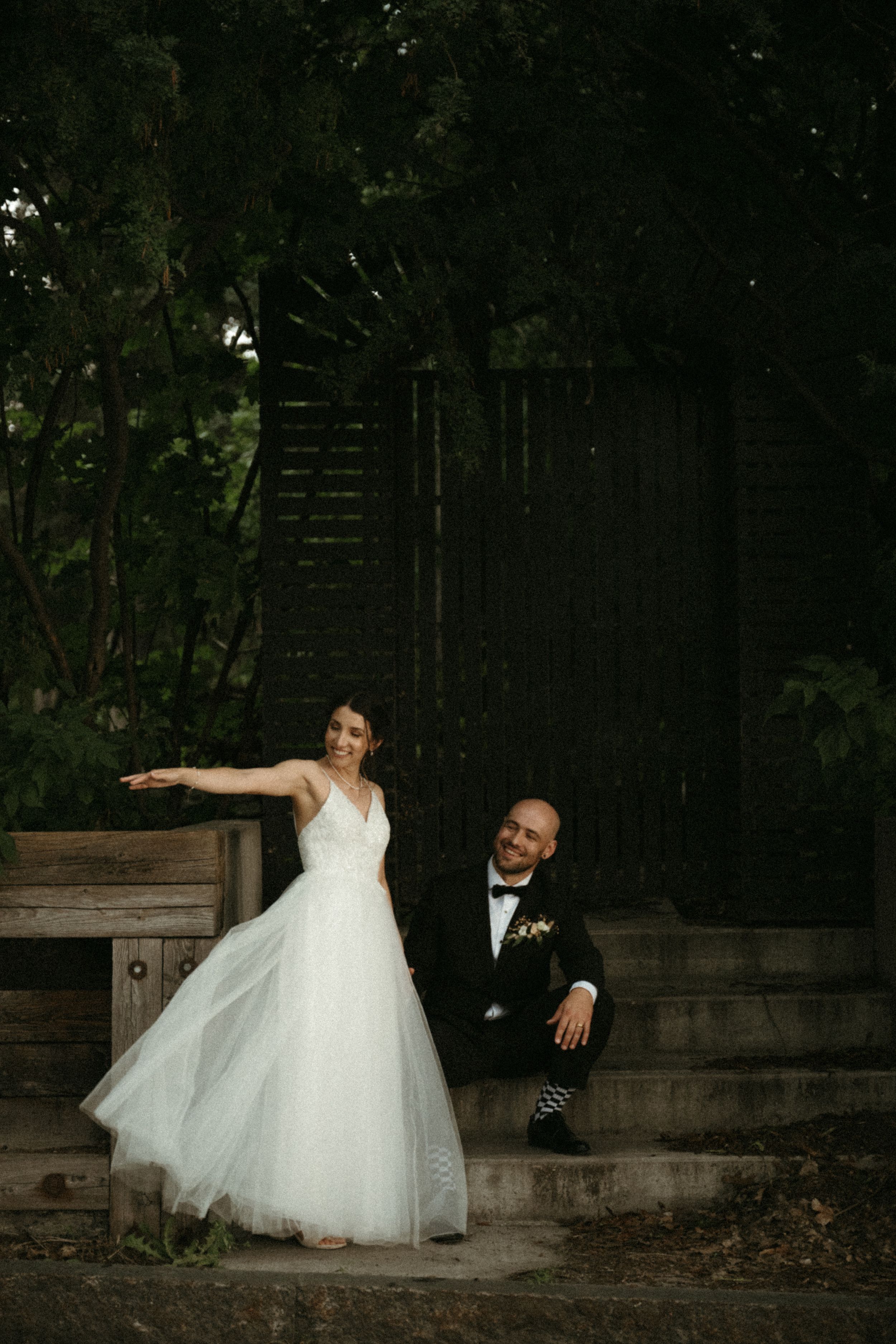 A wedding couple poses playfully on outdoor steps against a dark wooden wall.