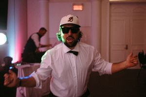 DJ in baseball cap and bowtie performing at wedding reception.