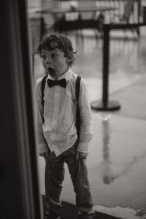 A young child in formal attire with a bow tie and suspenders stands in a hallway with a surprised expression.