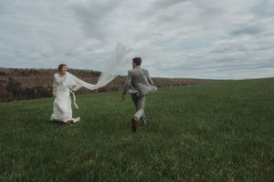 A playful bride and groom jumping together in a green field under cloudy skies.