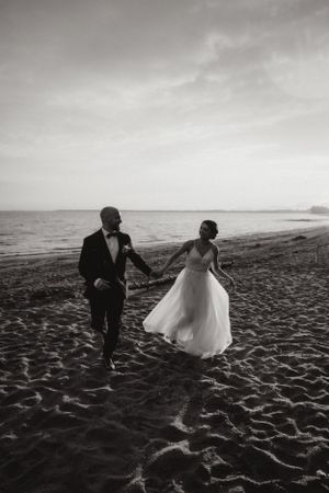A newlywed couple runs along the beach together in formal attire.