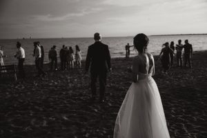 A couple in wedding attire stands on a beach with guests in the background.
