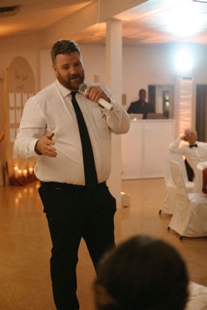 Un garçon d'honneur en chemise blanche et cravate noire parle dans un microphone lors d'une réception dans un lieu chaleureusement éclairé.