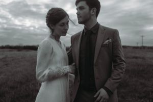 Black and white portrait of a wedding couple standing in an open field under cloudy skies.