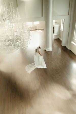 A bride in a white gown standing beneath an elegant crystal chandelier in a bright room.