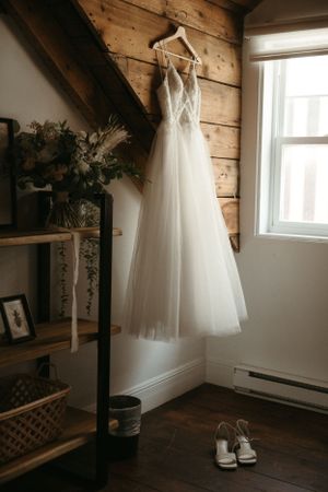 An elegant white wedding dress hangs on a wooden wall next to a window, with bridal shoes placed beneath it.