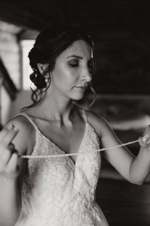 Portrait de mariage en noir et blanc montrant une mariée dans une robe en dentelle ajustant son collier dans un éclairage dramatique.