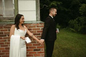 Couple holding papers while standing separately against brick wall during first look moment.