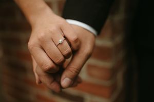 Close-up of two hands clasped together showing an engagement ring against a brick background.