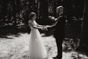 Un couple se tient la main lors d'une cérémonie de mariage intime en forêt dans un cadre boisé ensoleillé, capturé en noir et blanc.