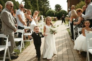 Young ring bearer in black suit walks down aisle during outdoor wedding ceremony.