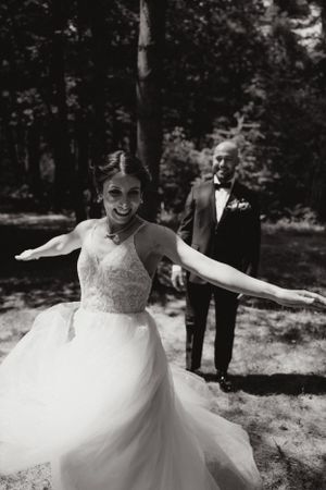 Photo en noir et blanc de la mariée en robe de tulle fluide. Elle danse joyeusement. Le marié admire sa femme.