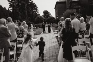 A wedding ceremony taking place outdoors with attendees seated in white chairs and pine trees in the background.
