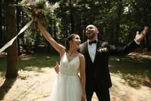 Newlyweds celebrate with raised arms under a rustic wedding arch in a woodland setting.