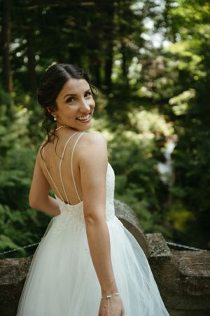 An elegant bride in a strappy white gown smiles over her shoulder in a forest setting.