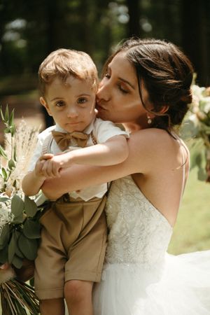 A tender moment between a bride in white and a small child in formal attire surrounded by greenery.