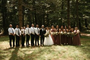 Wedding party stands in line against forest backdrop with bridesmaids in brown dresses and groomsmen in white shirts.