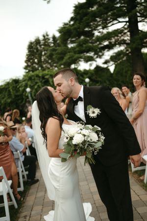 Romantic kiss between newlyweds on stone patio with wedding guests in background.