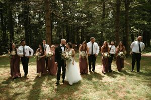 Wedding party laughs and poses casually in a sunlit forest clearing.