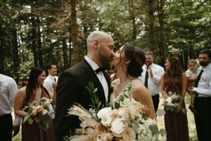 Newlyweds share intimate moment while holding white floral bouquets in wooded setting.