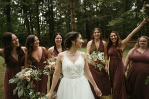 Bride and bridesmaids in burgundy dresses celebrate with bouquets in forest setting.