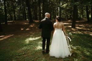 Couple walks hand in hand through sunlit forest in formal wedding attire.