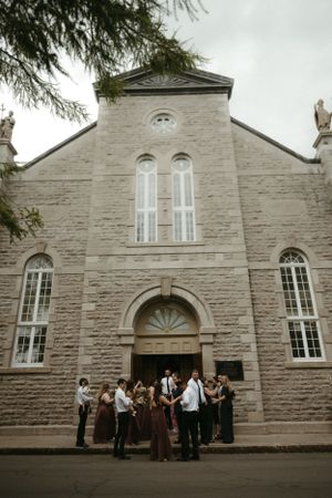 A wedding party gathers outside a historic stone church with tall arched windows and elegant architecture.