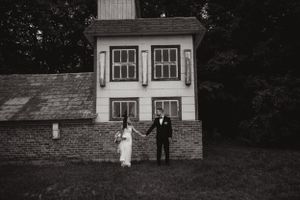 A minimalist wedding portrait of a couple standing against a small white house in black and white.