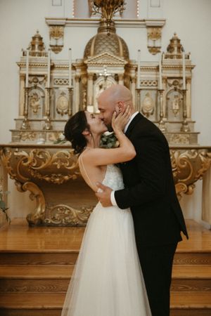A romantic first kiss moment during a wedding ceremony in front of an ornate golden altar.