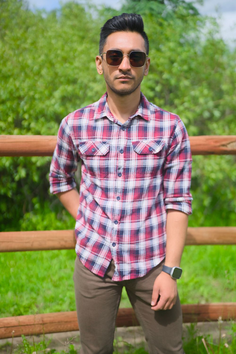 A person in a plaid red and white button-down shirt and tan pants poses against a wooden fence in a grassy outdoor setting.