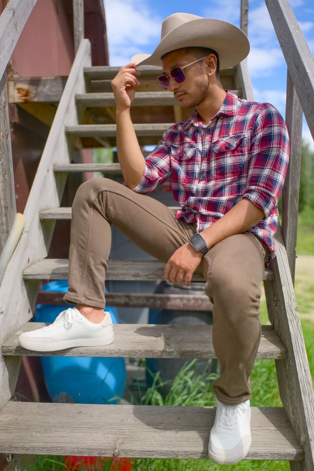 Person in khaki pants and plaid shirt with baseball cap sits casually on wooden stairs wearing white sneakers.
