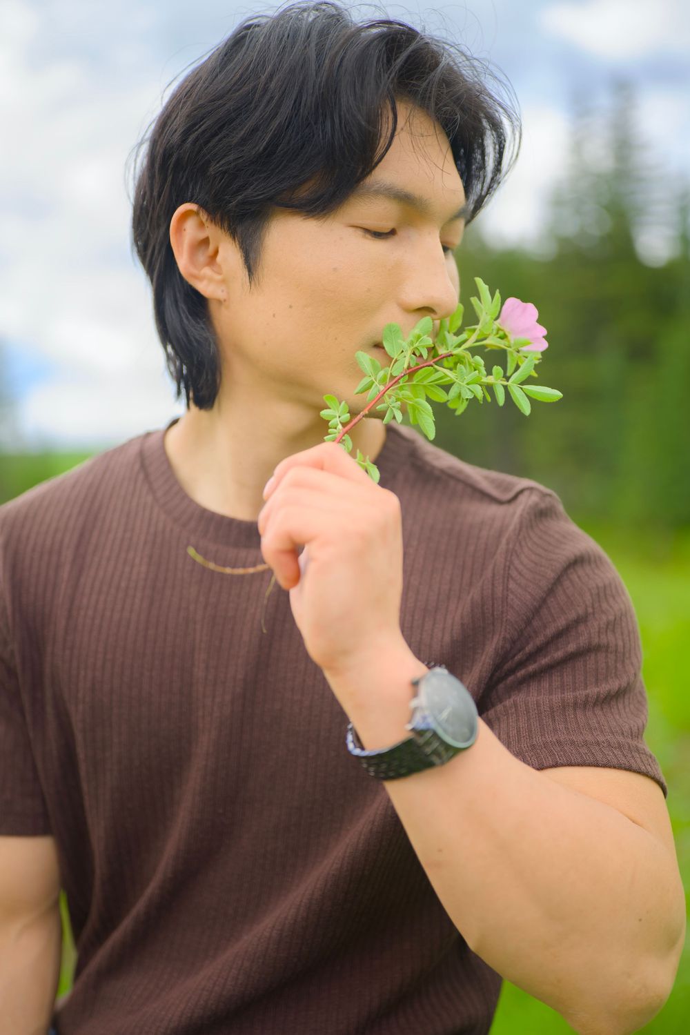 Person in a brown sweater smelling a delicate pink flower against a soft blurred nature background.