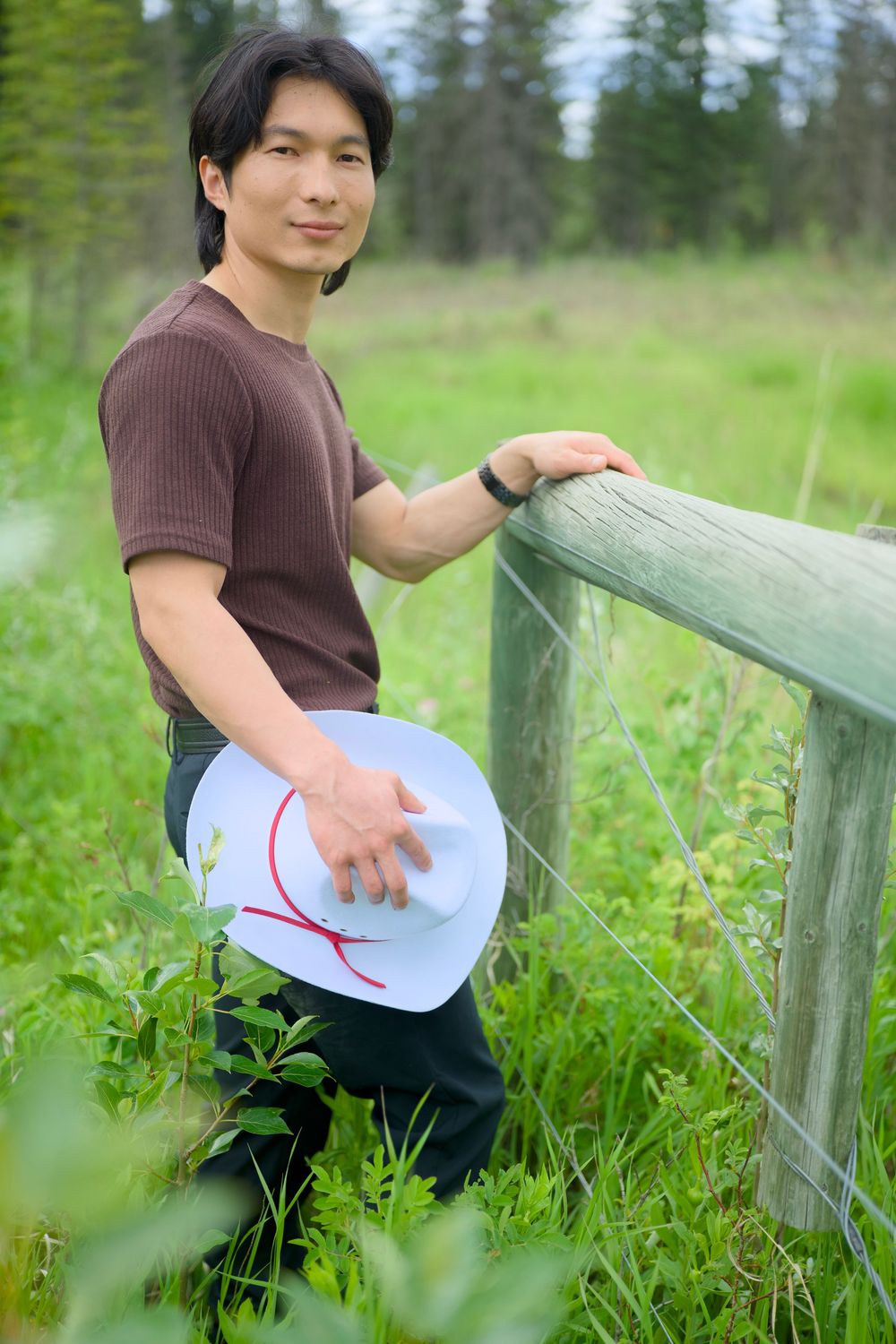Someone in a brown shirt holds a hat while leaning against a wooden fence in a grassy meadow with trees in background.
