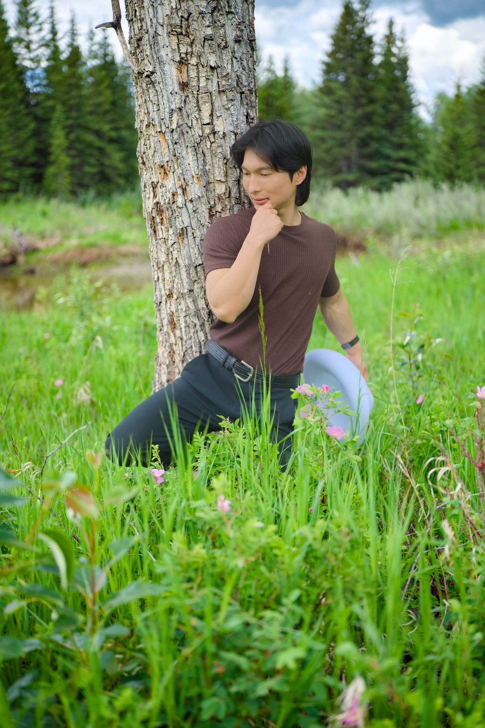 Someone in a brown shirt poses thoughtfully by a birch tree in a lush green meadow with evergreen forest in the background.