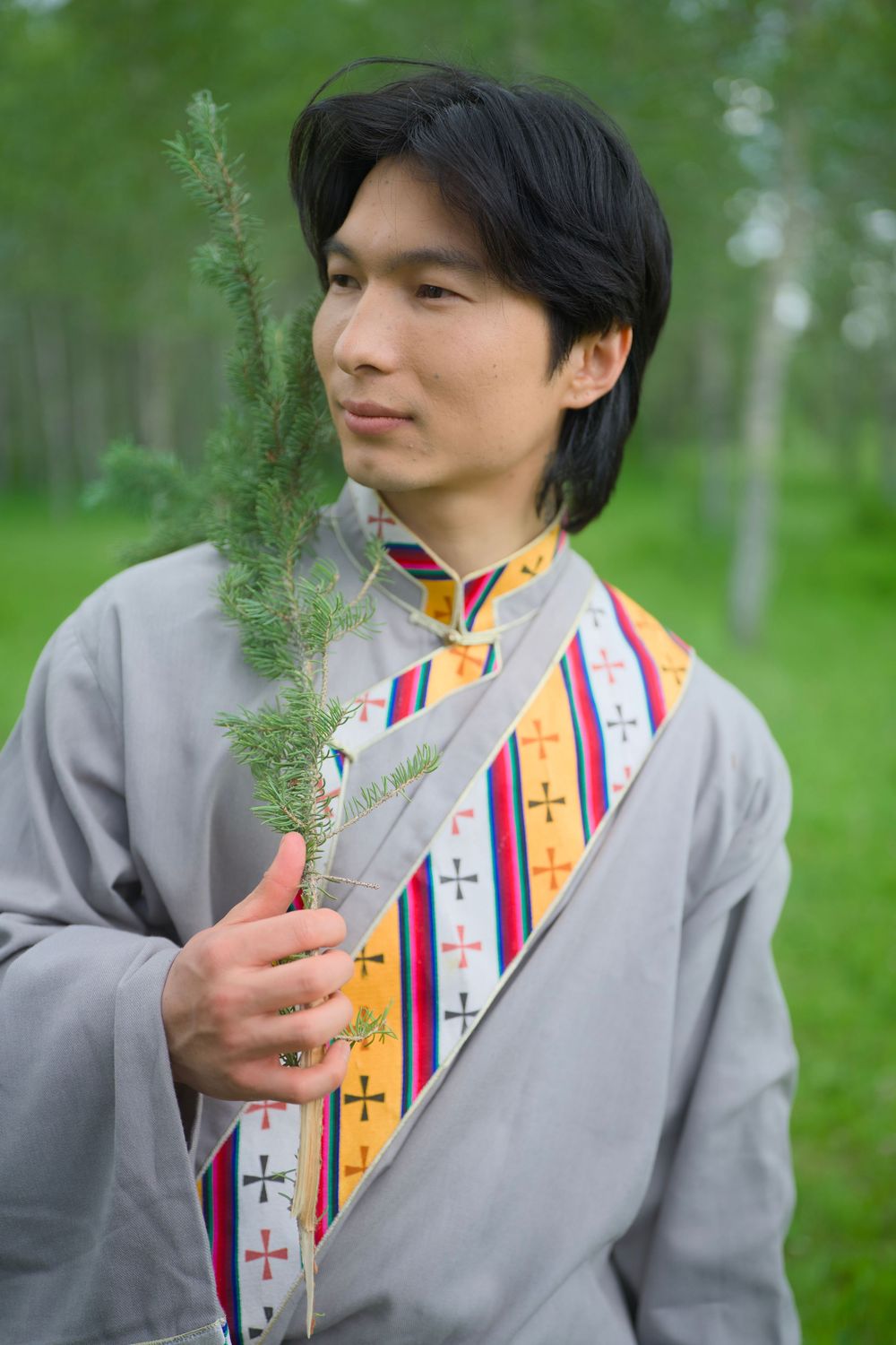 Person in gray clothing wearing a colorful striped stole poses thoughtfully in a lush green outdoor setting.