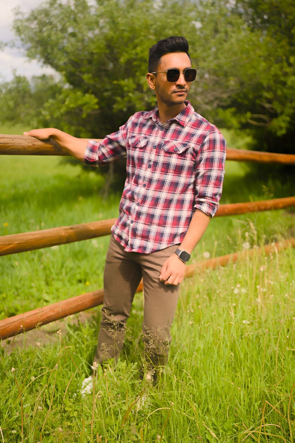 A person in a plaid shirt and sunglasses leans casually against a wooden fence in a green meadow on a sunny day.