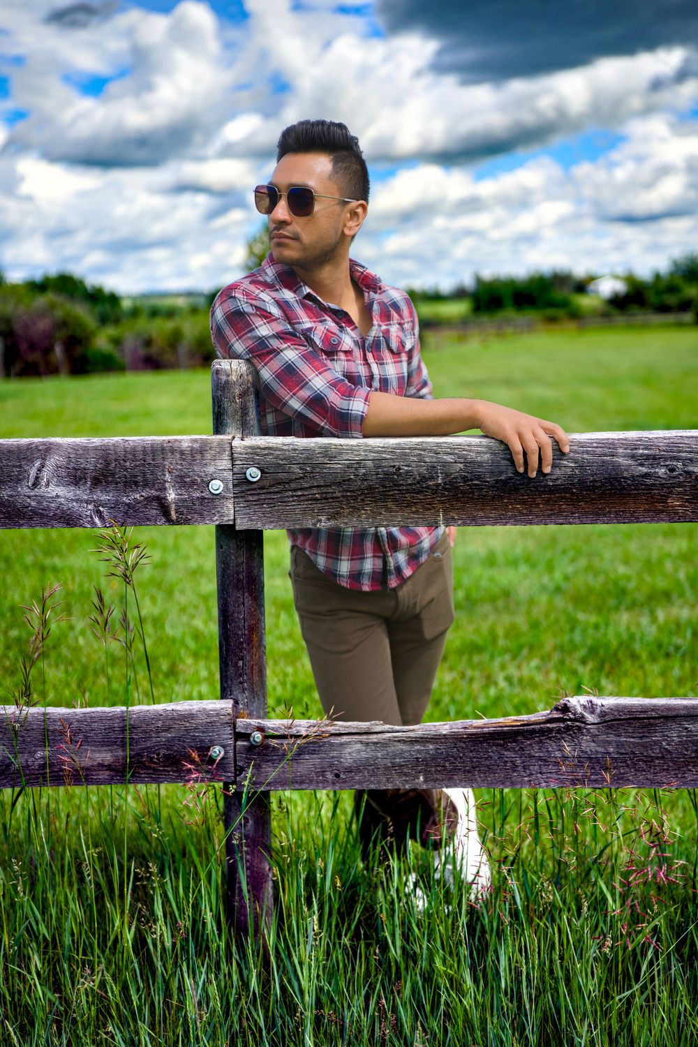 Someone in plaid shirt and sunglasses leans on rustic wooden fence surrounded by green meadow under dramatic clouds.