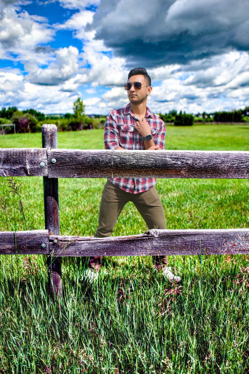 A person in sunglasses and plaid shirt leans on a rustic wooden fence with green fields and dramatic clouds in the background.