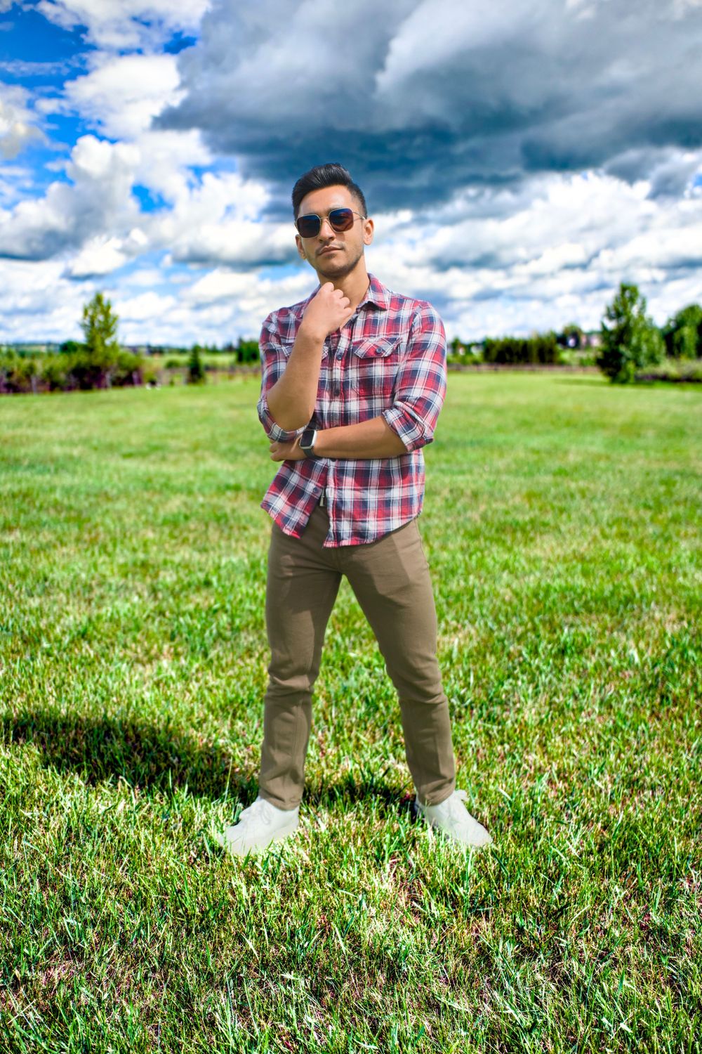 Someone in casual attire of plaid shirt and khaki pants poses stylishly on a grassy meadow under dramatic cloudy skies.