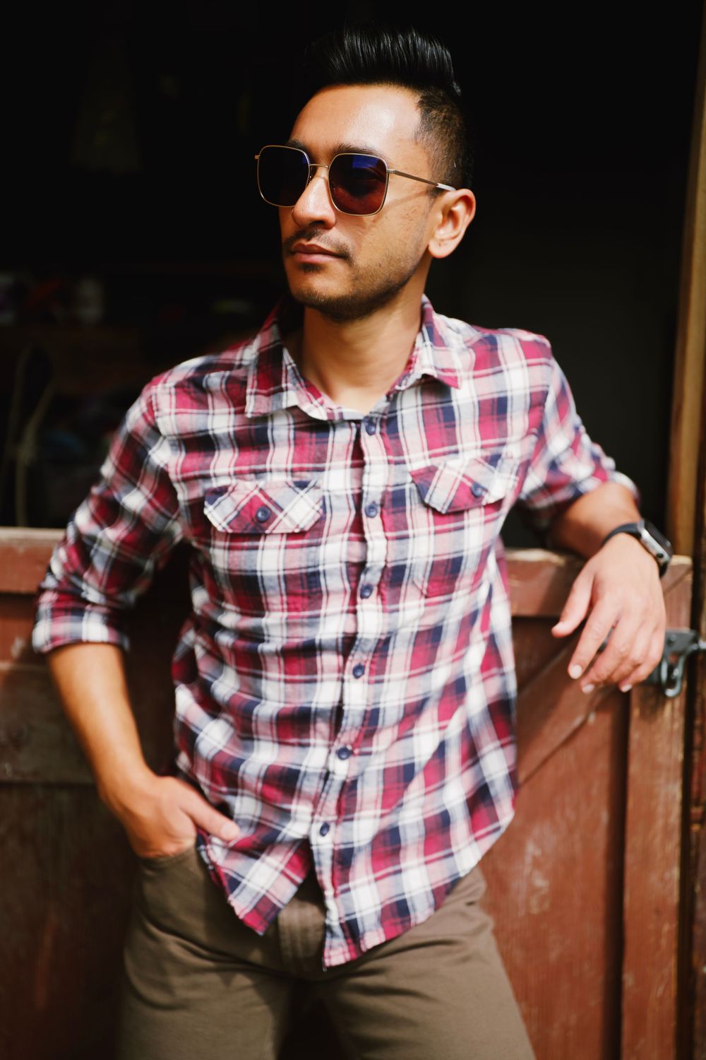 A person wearing aviator sunglasses and a red plaid button-up shirt leans against a wooden wall in casual outdoor setting.